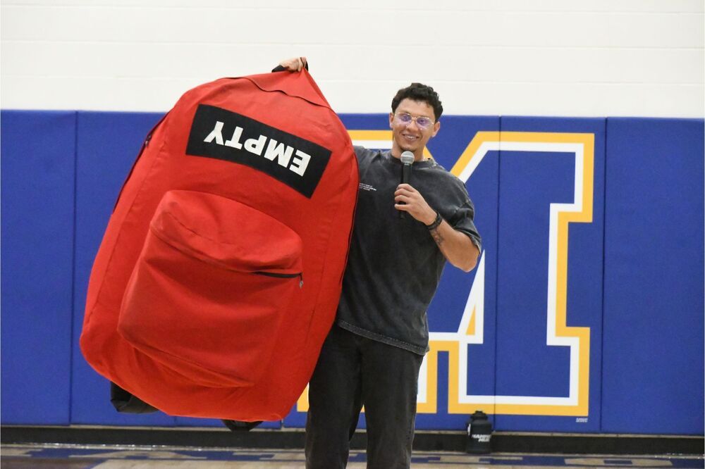 Guest Speaker holding up a giant backpack as he speaks to an assembly