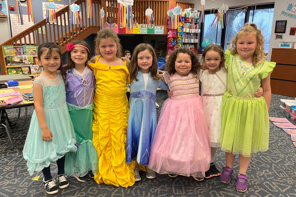 7 kindergarten students dressed in disney princess dresses in a classroom.