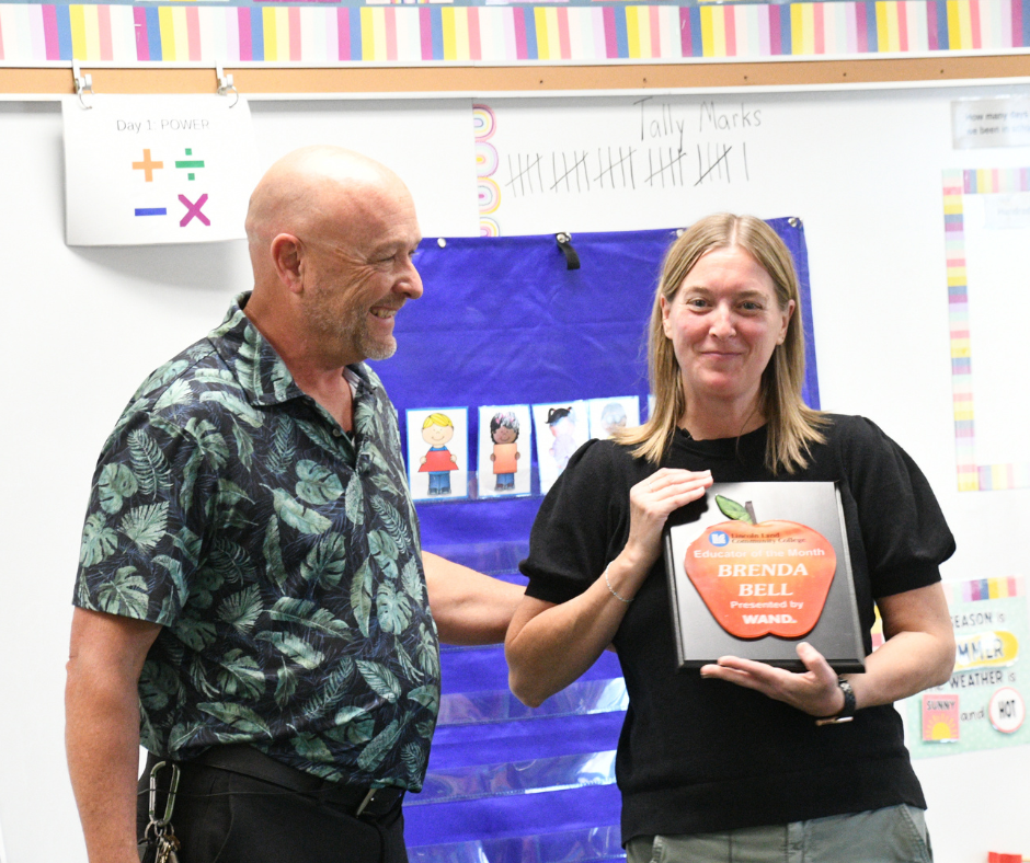 Williams Principal Mike Shaffer handing the WAND Educator of the Month plaque to first grade teacher Brenda Bell in front of her classroom