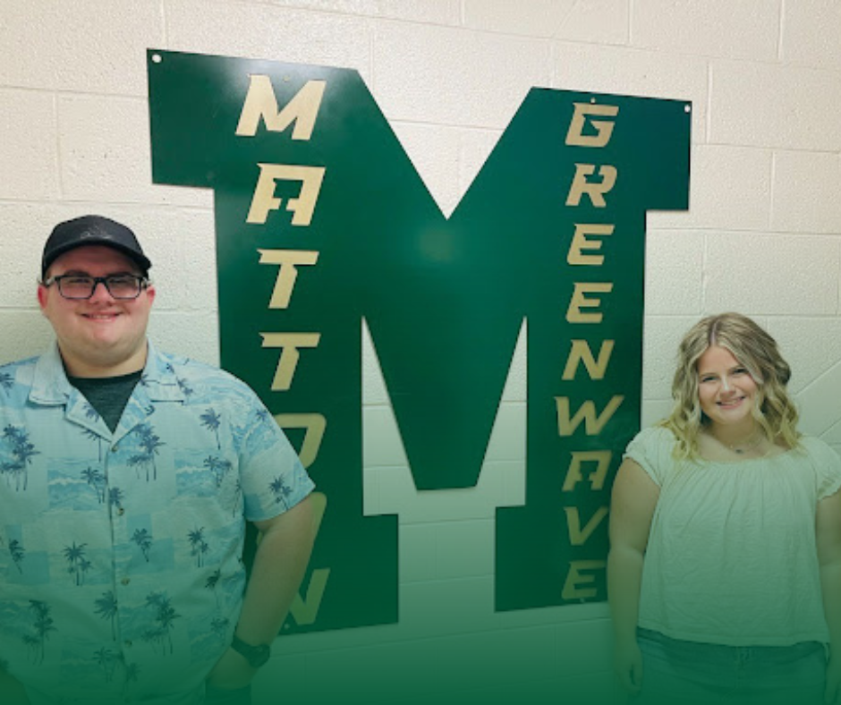 Two MHS students smiling next to a Mattoon Greenwave logo hanging in a hallway. They are pictured for being the Mattoon Rotary Students of the Month