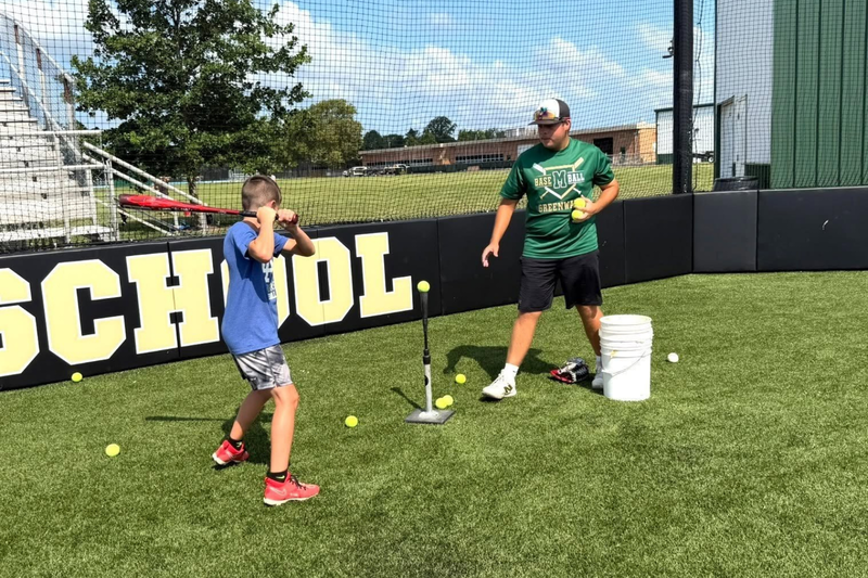MHS youth summer camp. Kid hitting a baseball bat with a coach nearby.