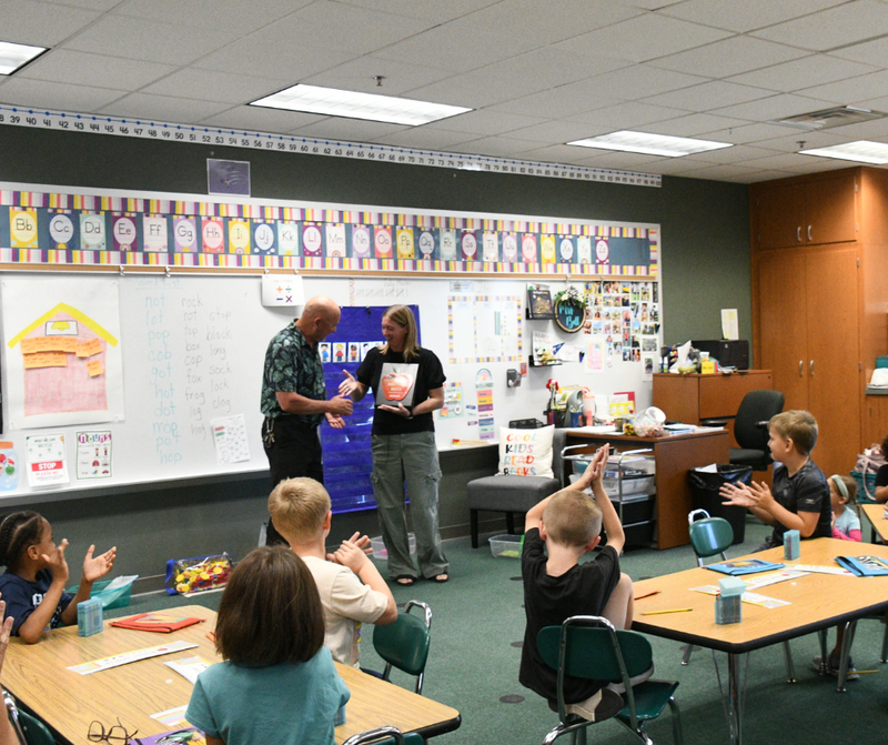 Williams Principal Mike Shaffer handing the WAND Educator of the Month plaque to first grade teacher Brenda Bell in front of her classroom. Students are clapping at their desks