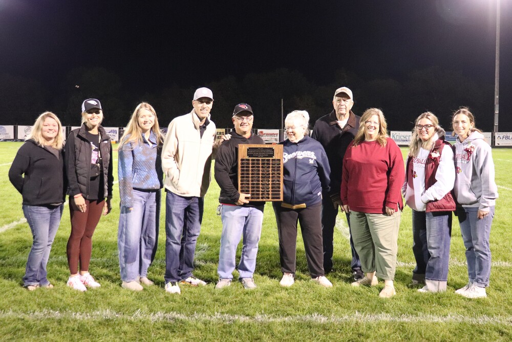 2026 Katie Award Winner Brian Thom and family and the Mayville Athletic Booster Club Board smiling on football field at halftime award presentation