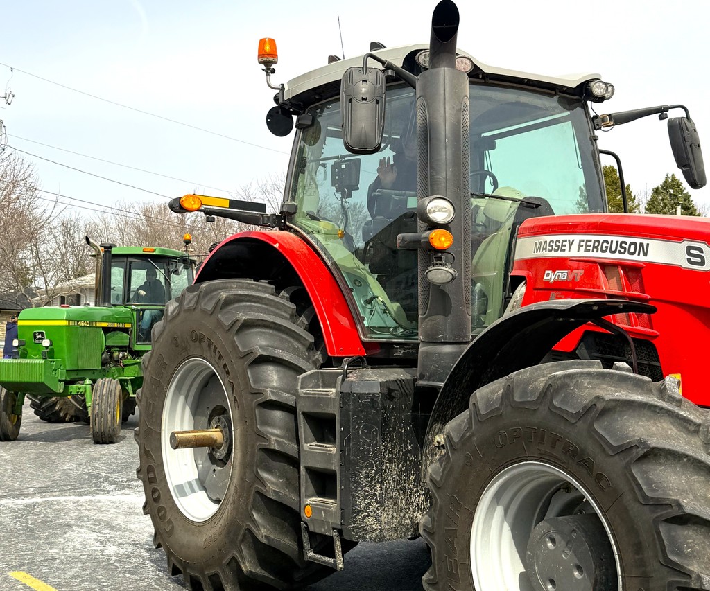 High School student waiving from driver's seat of red Massey Ferguson tractor, followed by green John Deere tractor driven by another student driver.