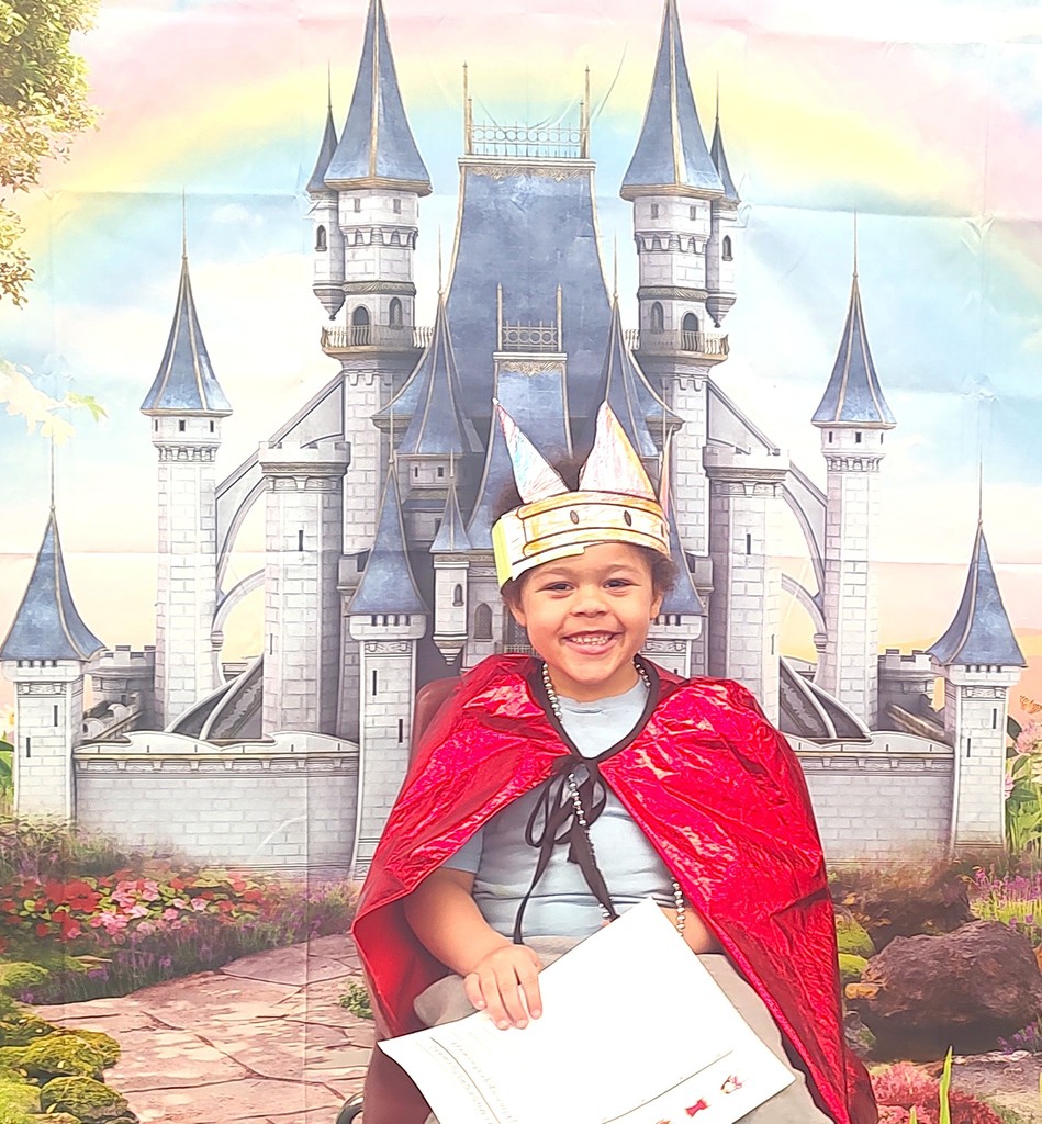 child dressed in royal crown and red cape smiling in front of backdrop with castle