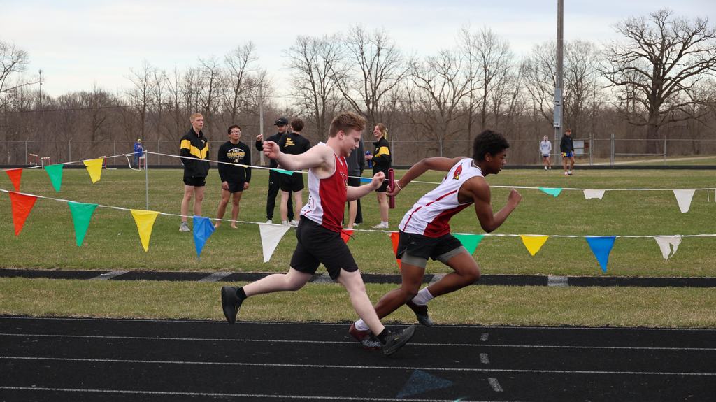Mayville Track team Berry Invite runners on track with green grass infield, multi-colored flags and other team in background