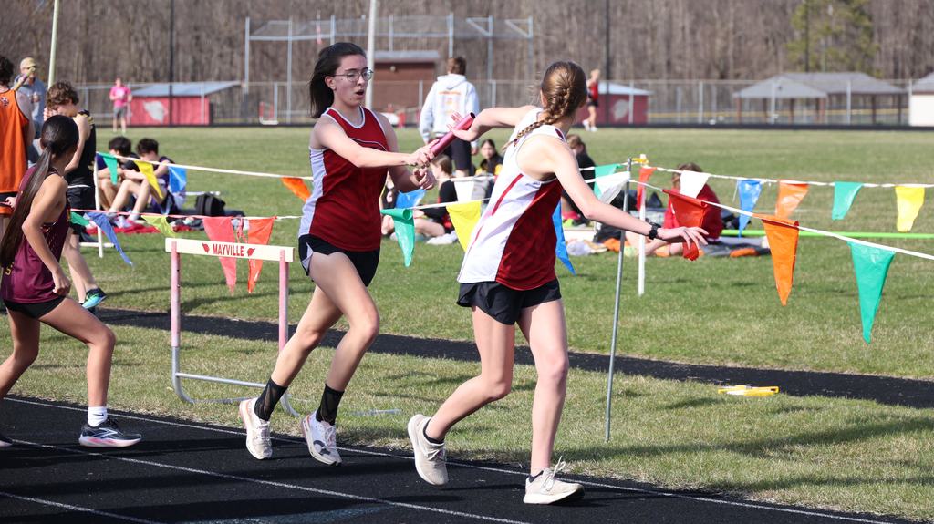 Mayville Track team Berry Invite runners on track doing stick hand-off green grass infield, multi-colored flags and other track participants in background