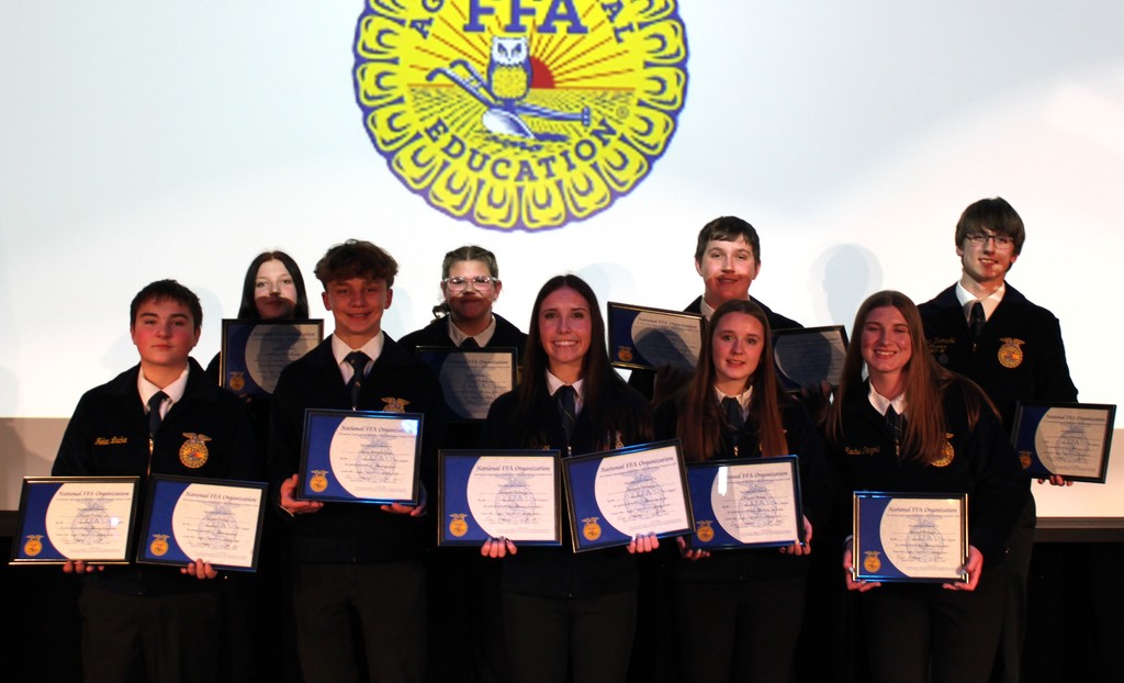 FFA students smiling for photo on auditorium stage with degree certificates and FFA emblem in the background