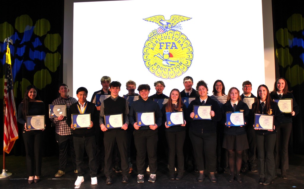 FFA students smiling for photo on auditorium stage with degree certificates and FFA emblem in the background