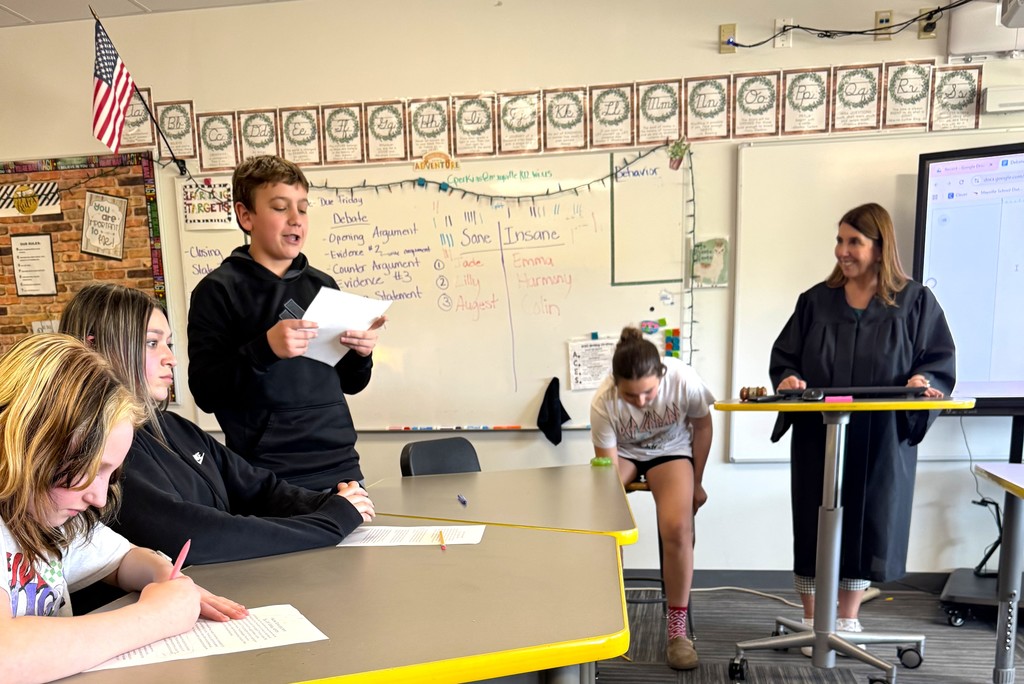 7th graders in Mrs. Perkins' classroom participating in a mock trial as part of their ELA class, reading arguments they've prepared. Mrs. Perkins is dressed in a judge robe with a wooden gavel. 