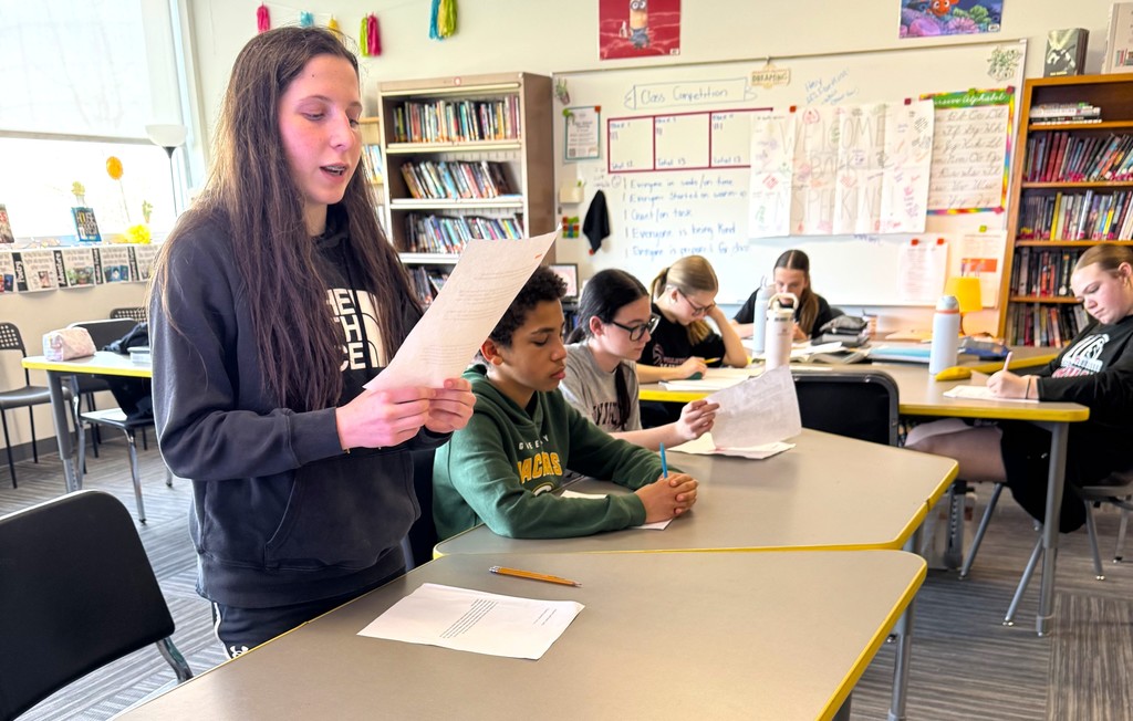 7th graders in Mrs. Perkins' classroom participating in a mock trial as part of their ELA class, reading arguments they've prepared. Mrs. Perkins is dressed in a judge robe with a wooden gavel. 