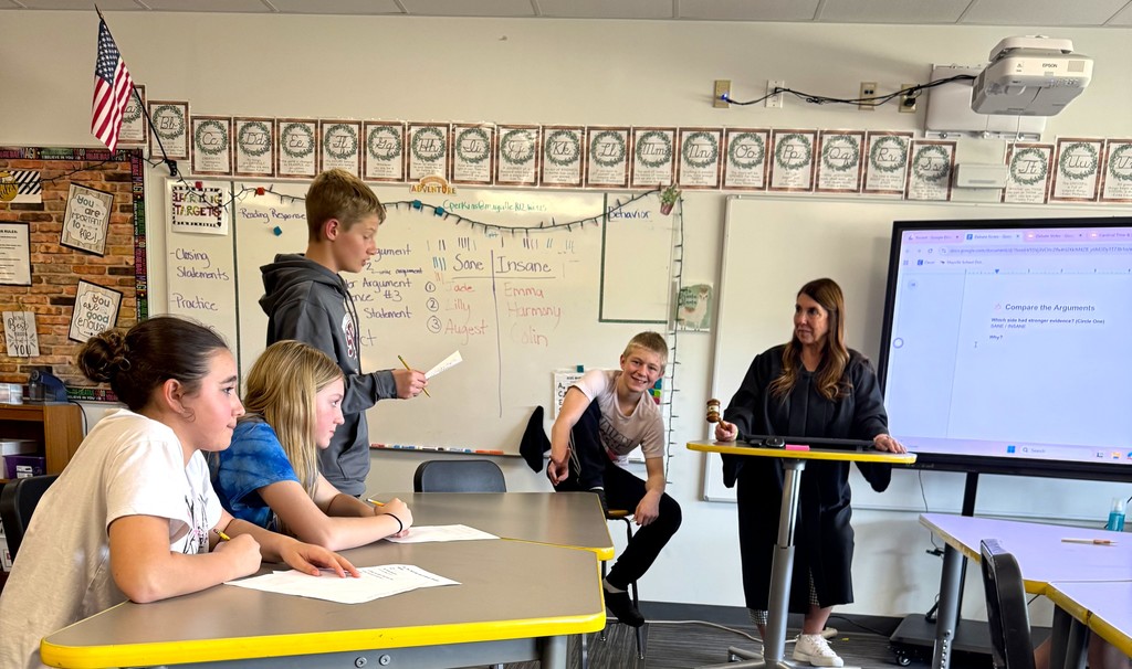 7th graders in Mrs. Perkins' classroom participating in a mock trial as part of their ELA class, reading arguments they've prepared. Mrs. Perkins is dressed in a judge robe with a wooden gavel. 
