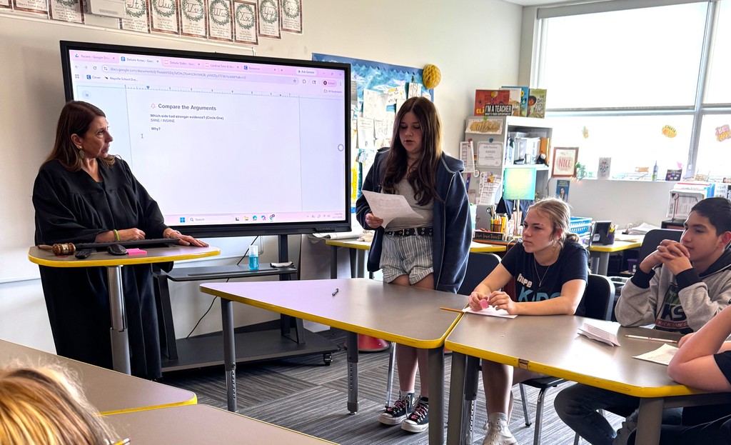 7th graders in Mrs. Perkins' classroom participating in a mock trial as part of their ELA class, reading arguments they've prepared. Mrs. Perkins is dressed in a judge robe with a wooden gavel. 