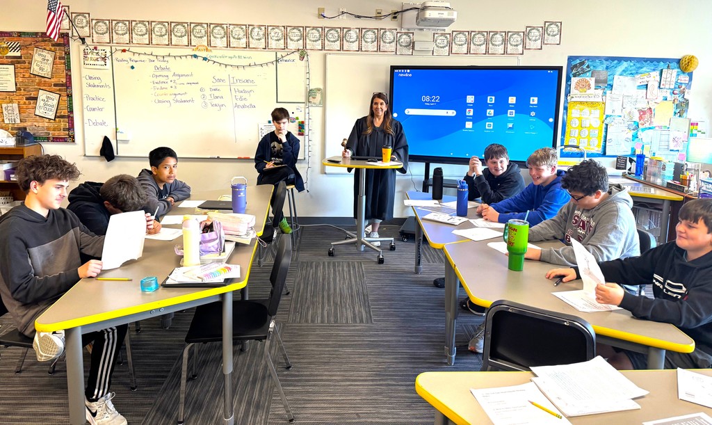 7th graders in Mrs. Perkins' classroom participating in a mock trial as part of their ELA class, reading arguments they've prepared. Mrs. Perkins is dressed in a judge robe with a wooden gavel. 
