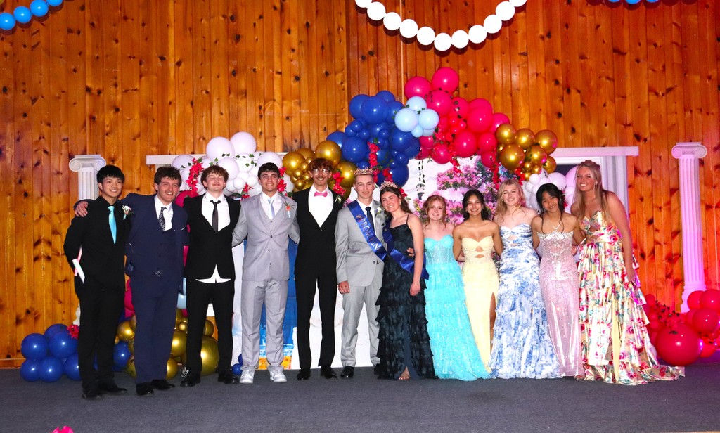 MHS Junior Prom Court photo with boys in suits and girls in formal dresses and wood of Mayville Pavilion dance hall in background along with rainbow balloons and Greek pillars