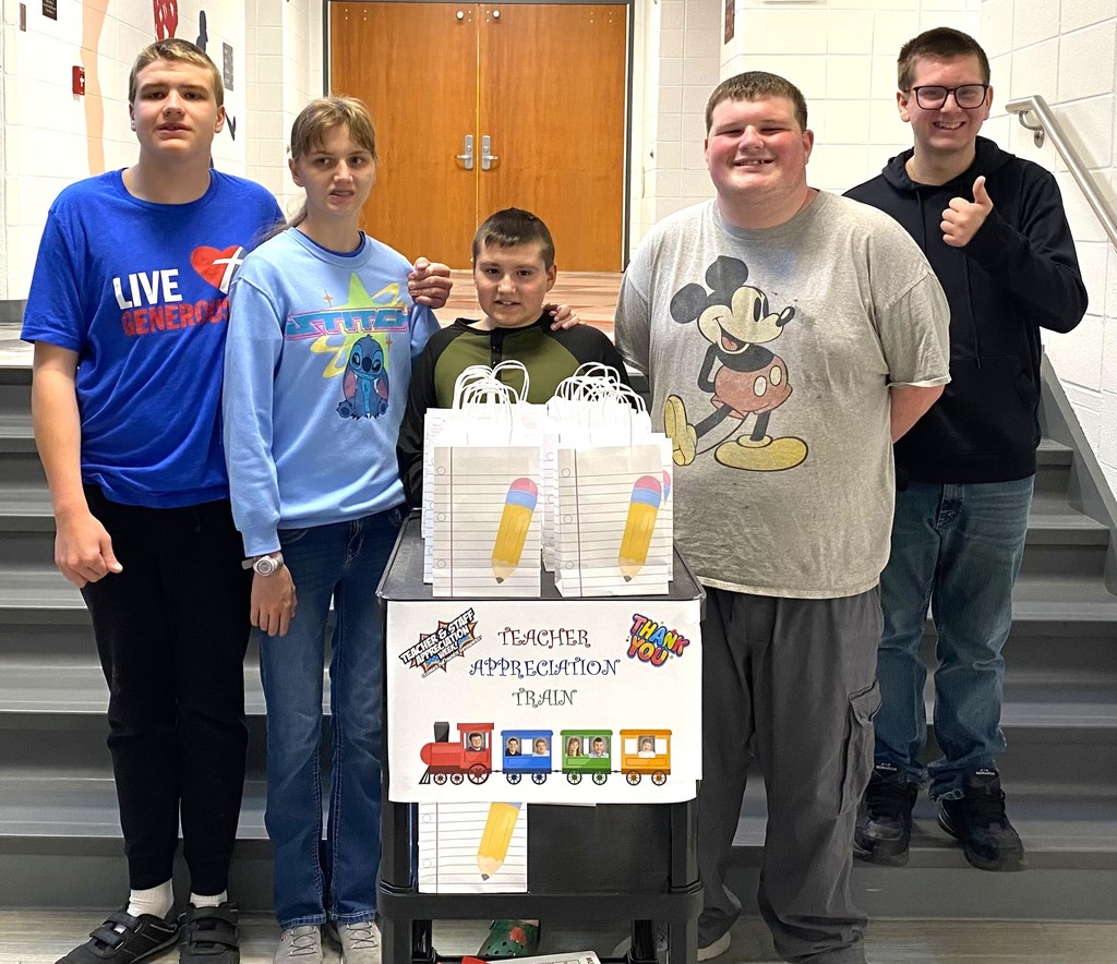 Photo of students smiling and standing in stairway with a cart full of teacher appreciation gifts and a sign that says Teacher Appreciation Train