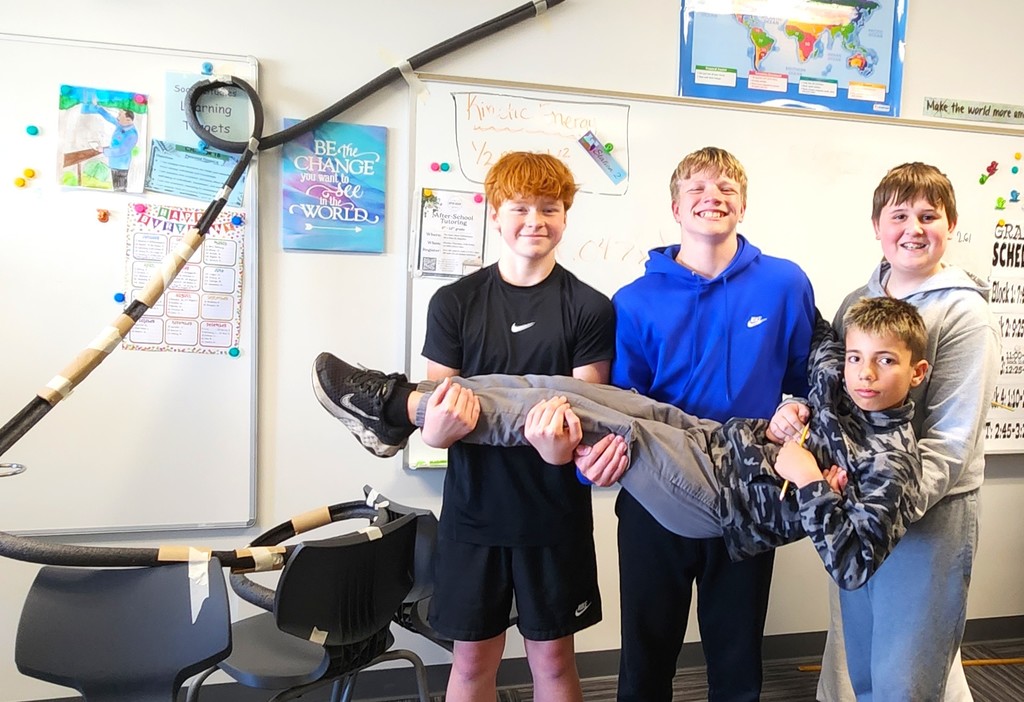 Students in classroom smiling next to their rollercoaster creations made of flexible rubber tubing, paper toilet and towel rolls, and adhered to the wall with tape