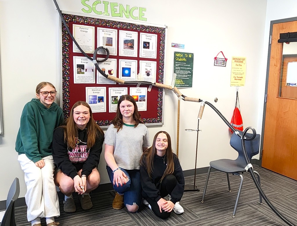 Students in classroom smiling next to their rollercoaster creations made of flexible rubber tubing, paper toilet and towel rolls, and adhered to the wall with tape