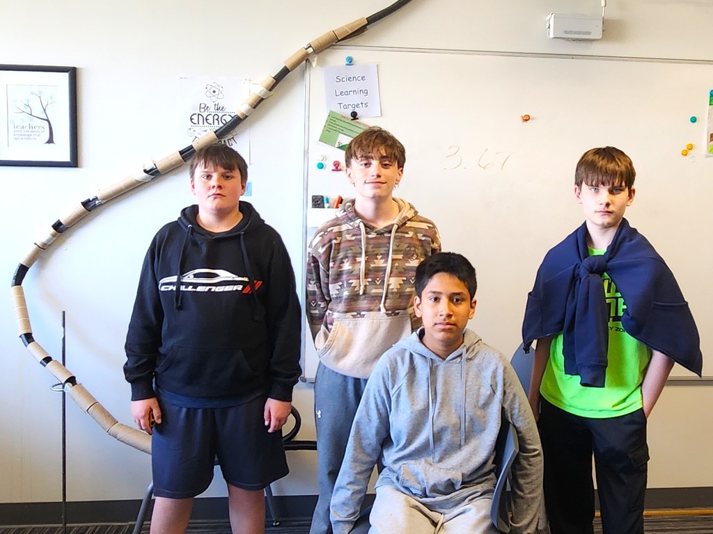 Students in classroom smiling next to their rollercoaster creations made of flexible rubber tubing, paper toilet and towel rolls, and adhered to the wall with tape