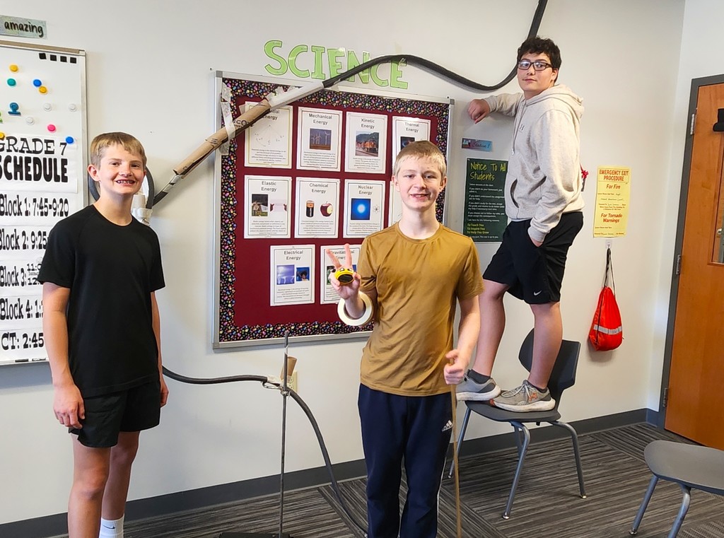 Students in classroom smiling next to their rollercoaster creations made of flexible rubber tubing, paper toilet and towel rolls, and adhered to the wall with tape
