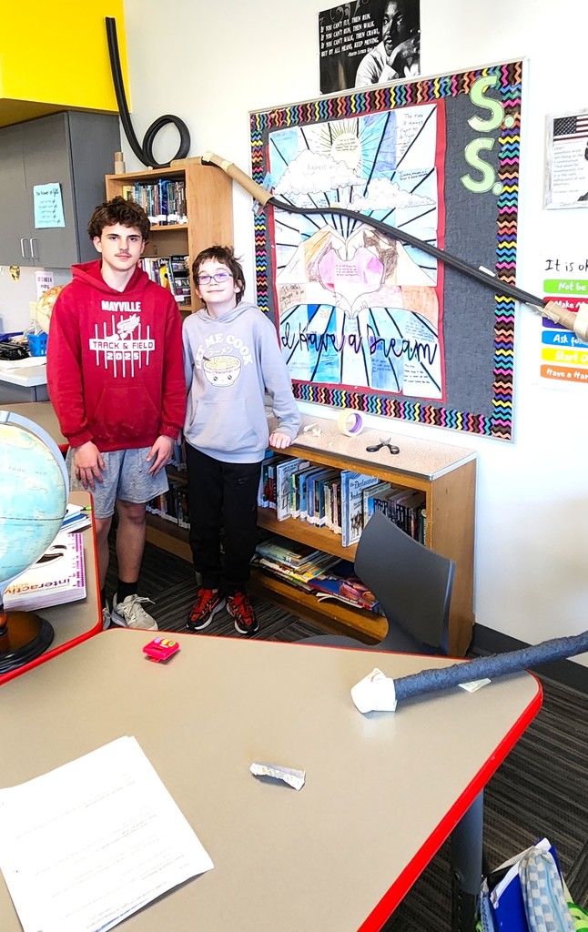 Students in classroom smiling next to their rollercoaster creations made of flexible rubber tubing, paper toilet and towel rolls, and adhered to the wall with tape