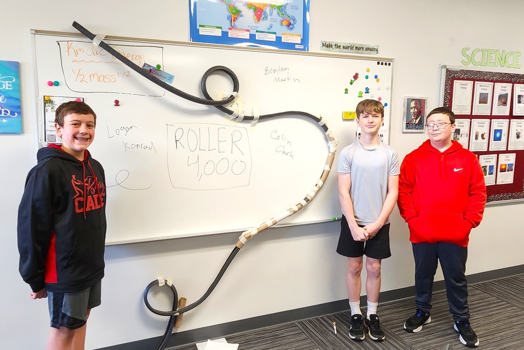 Students in classroom smiling next to their rollercoaster creations made of flexible rubber tubing, paper toilet and towel rolls, and adhered to the wall with tape