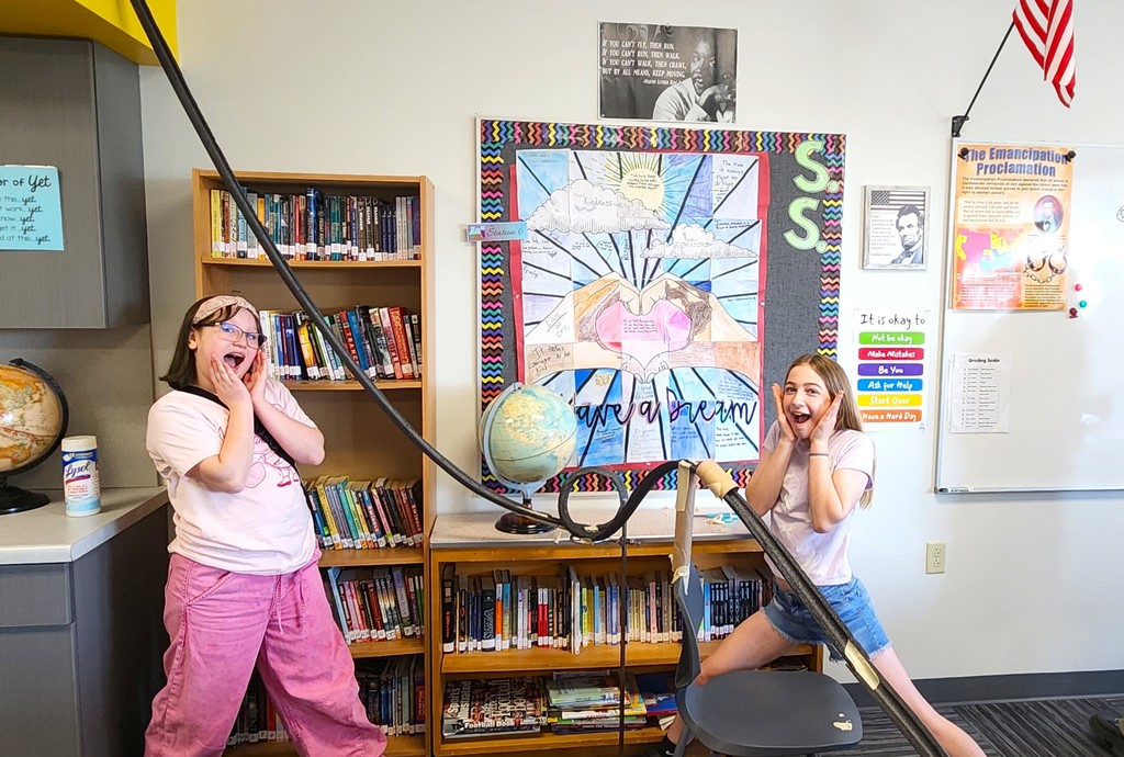 Students in classroom smiling next to their rollercoaster creations made of flexible rubber tubing, paper toilet and towel rolls, and adhered to the wall with tape