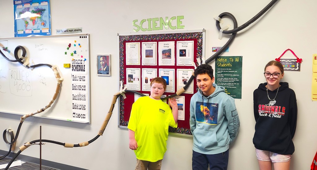 Students in classroom smiling next to their rollercoaster creations made of flexible rubber tubing, paper toilet and towel rolls, and adhered to the wall with tape
