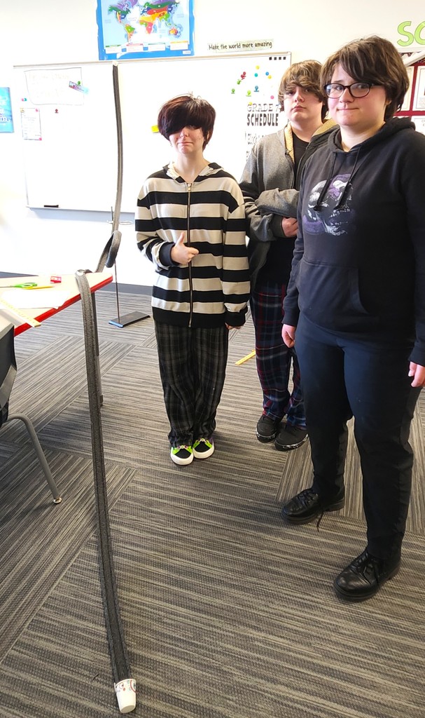 Students in classroom smiling next to their rollercoaster creations made of flexible rubber tubing, paper toilet and towel rolls, and adhered to the wall with tape