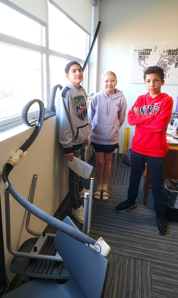 Students in classroom smiling next to their rollercoaster creations made of flexible rubber tubing, paper toilet and towel rolls, and adhered to the wall with tape