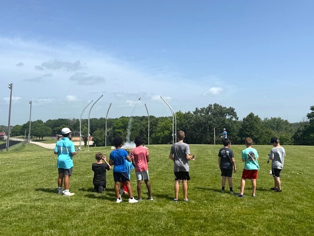 Summer school students out on lawn near school launching rockets and smiling