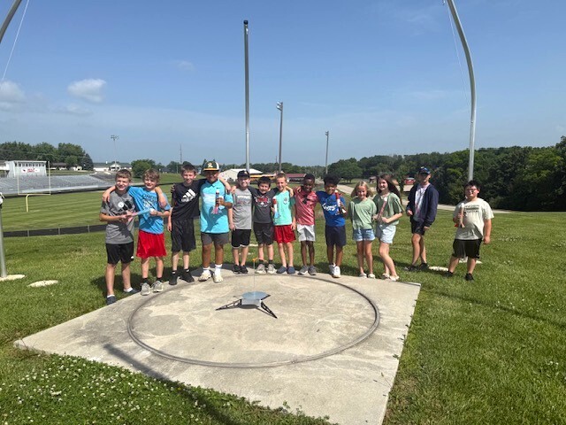Summer school students out on lawn near school launching rockets and smiling