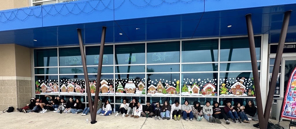 “Students sit in a long row outside a building decorated with colorful gingerbread house window murals.”