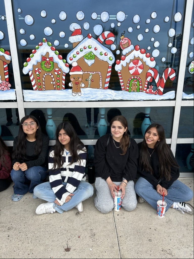 “Students sit in a long row outside a building decorated with colorful gingerbread house window murals.”