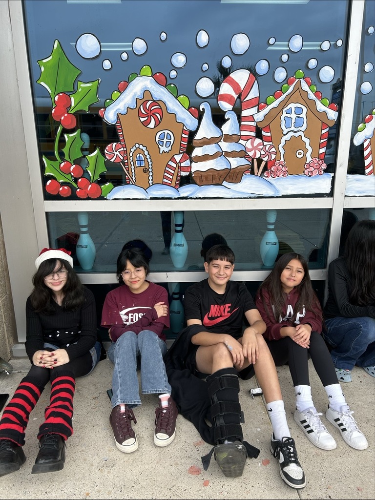 “Students sit in a long row outside a building decorated with colorful gingerbread house window murals.”