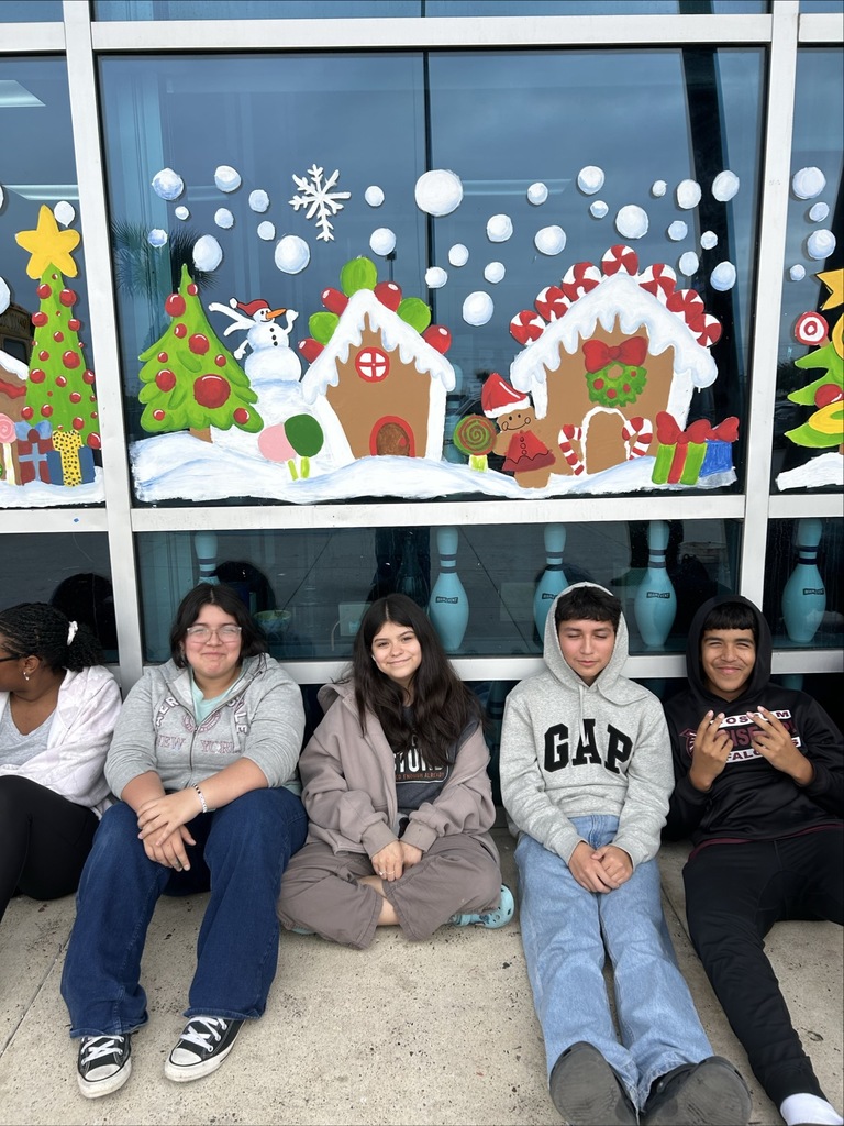 “Students sit in a long row outside a building decorated with colorful gingerbread house window murals.”