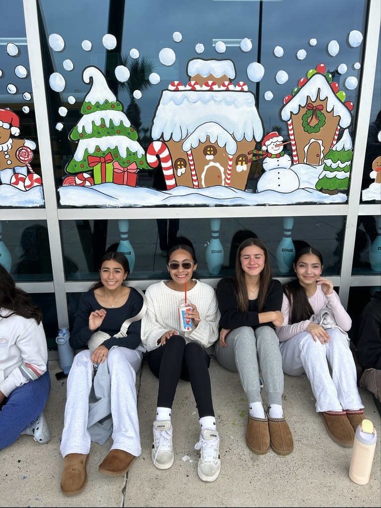 “Students sit in a long row outside a building decorated with colorful gingerbread house window murals.”