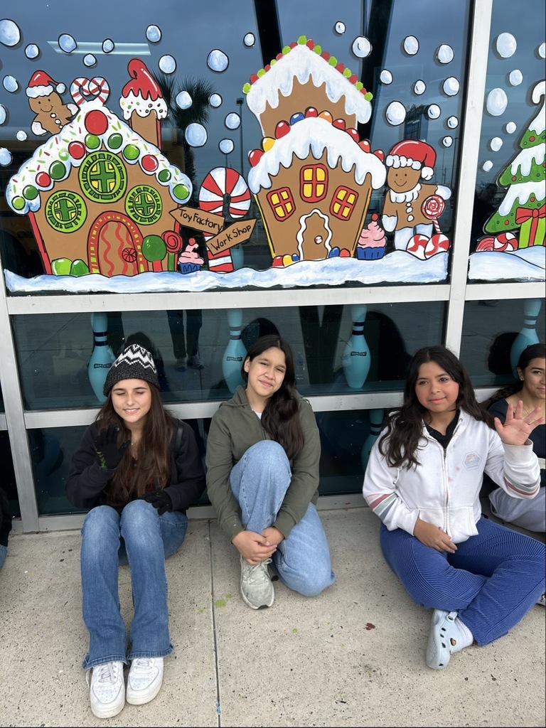 “Students sit in a long row outside a building decorated with colorful gingerbread house window murals.”