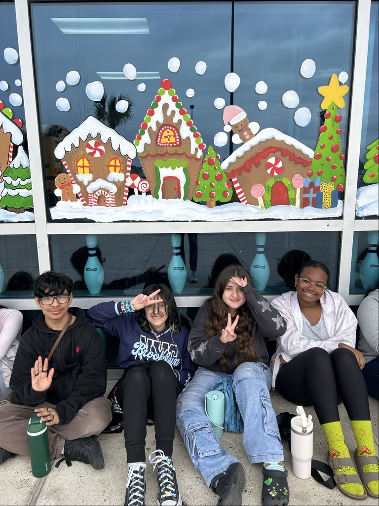 “Students sit in a long row outside a building decorated with colorful gingerbread house window murals.”