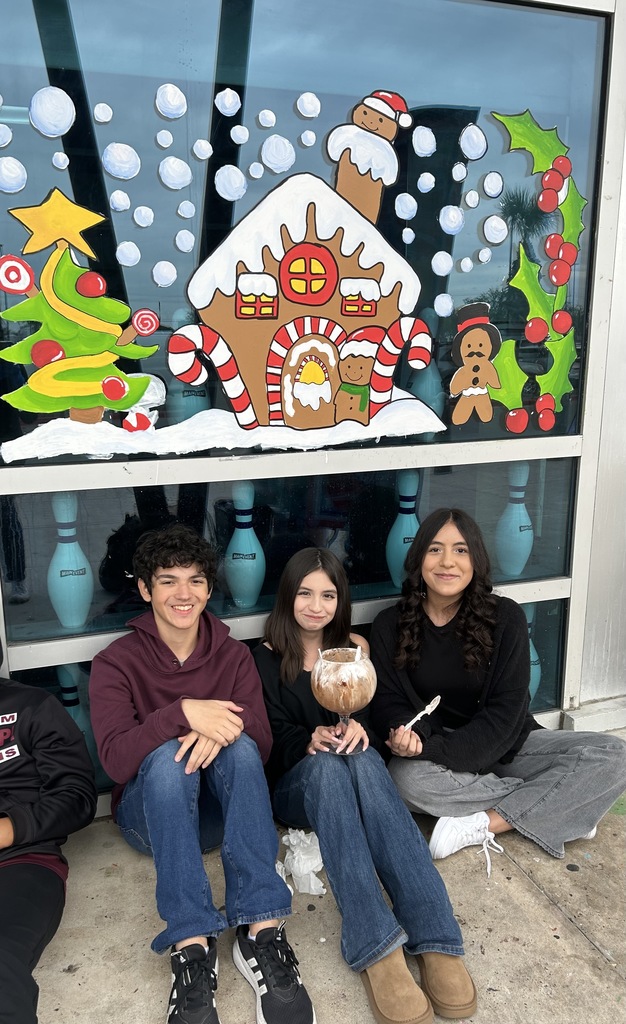 “Students sit in a long row outside a building decorated with colorful gingerbread house window murals.”