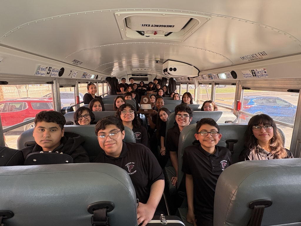 Fossum students seated on a school bus smiling toward the camera, with one student in the center holding a UIL plaque trophy. The group appears excited and proud as they travel together, wearing matching black Fossum shirts.