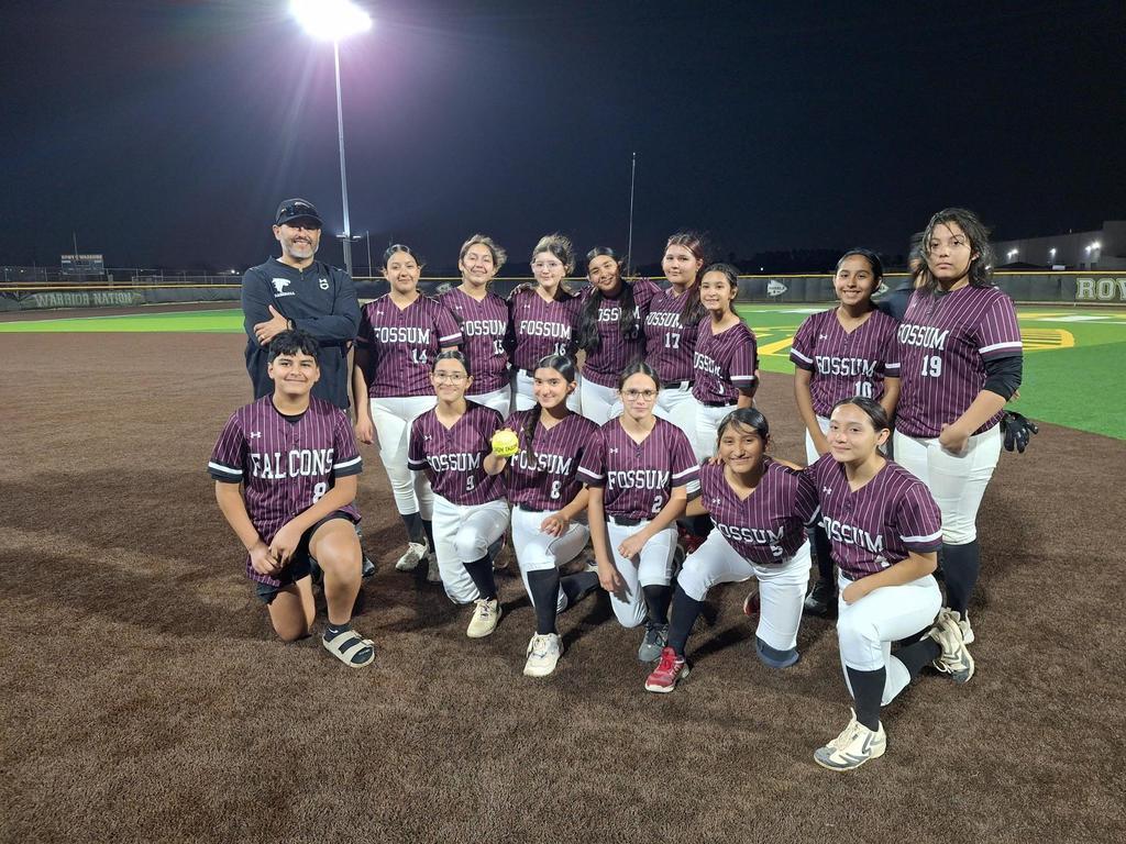 Fossum Falcons softball team posing together on the field at night under stadium lights. Players wear maroon Fossum jerseys with white pants, and a coach stands behind them. One player kneeling in front holds a yellow softball as the team smiles proudly after the game.