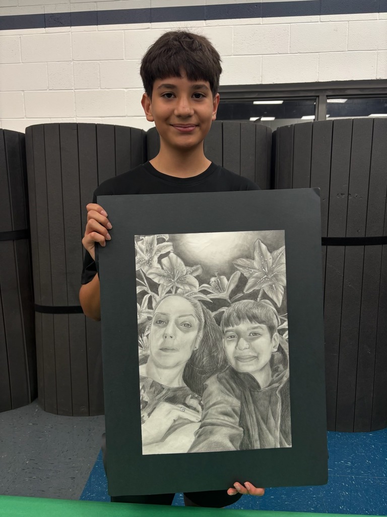 A middle school student stands indoors holding a large framed pencil drawing. The artwork is a detailed black-and-white portrait of two people surrounded by flowers, showing strong shading and realistic features. The student smiles proudly while presenting the piece, standing in front of a neutral wall with folded cafeteria tables behind them.