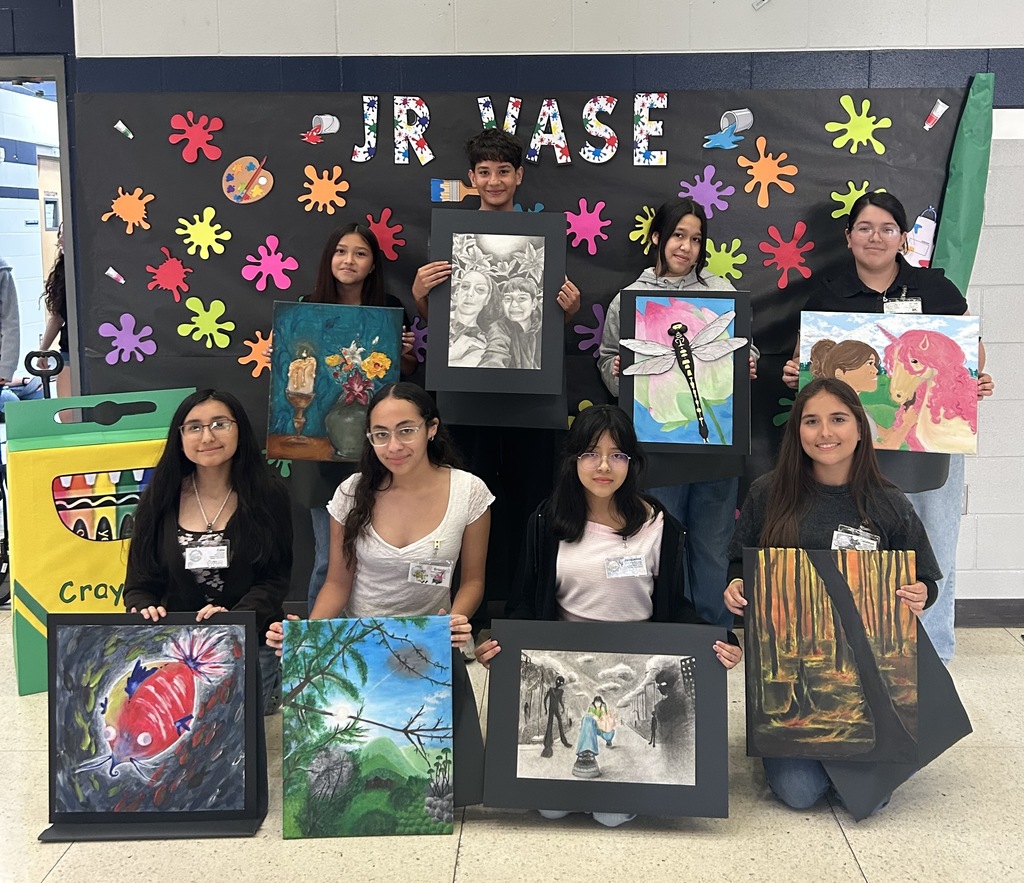 A group of middle school students pose together in a school hallway in front of a colorful “JR VASE” display decorated with paint splatters. Each student holds their artwork, including paintings of landscapes, still life, animals, and a detailed pencil drawing. The group smiles proudly, showcasing their creativity and achievements in art.
