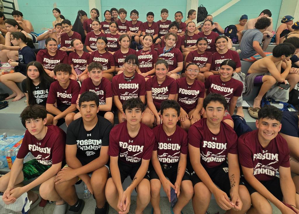 A large group of Fossum Middle School students sit together on indoor pool bleachers wearing matching maroon “Fossum Swimming” shirts. The team includes boys and girls smiling and posing for a group photo, while other students in swim attire sit in the background. The setting appears to be a swim meet, with towels, gear, and water bottles visible around them.