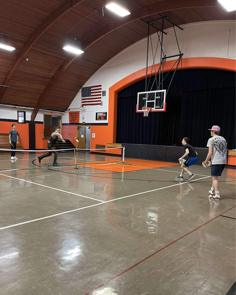 A deputy sheriff playing pickleball in a high school gym with three teenage boys.