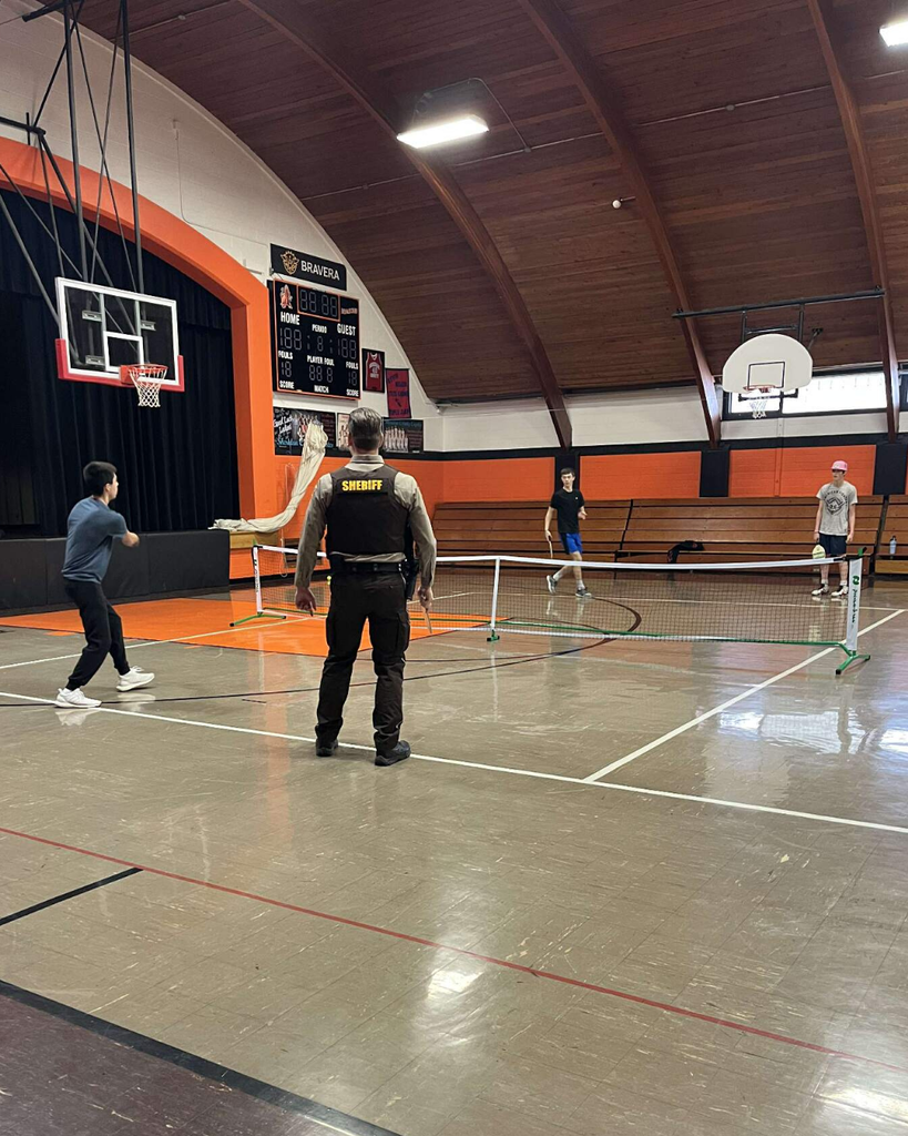 A deputy sheriff playing pickleball in a high school gym with three teenage boys.