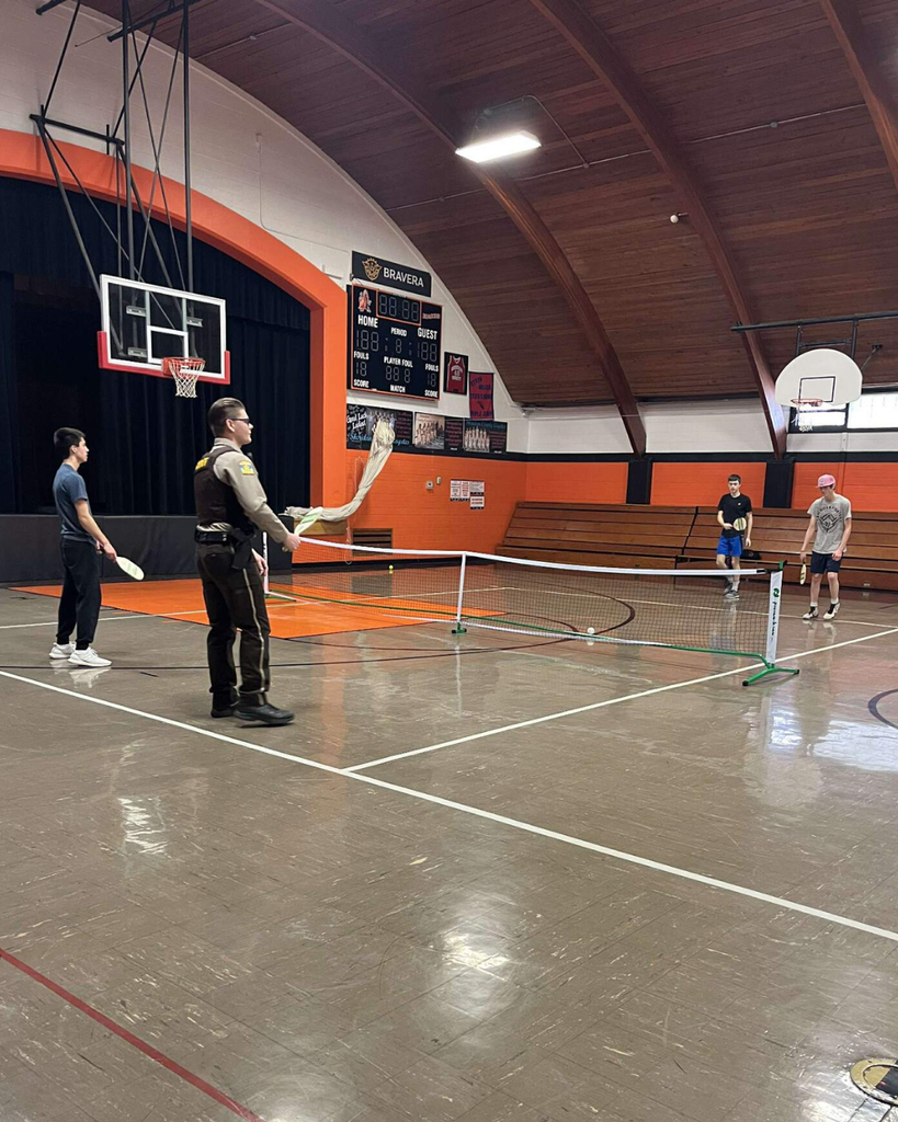A deputy sheriff playing pickleball in a high school gym with three teenage boys.