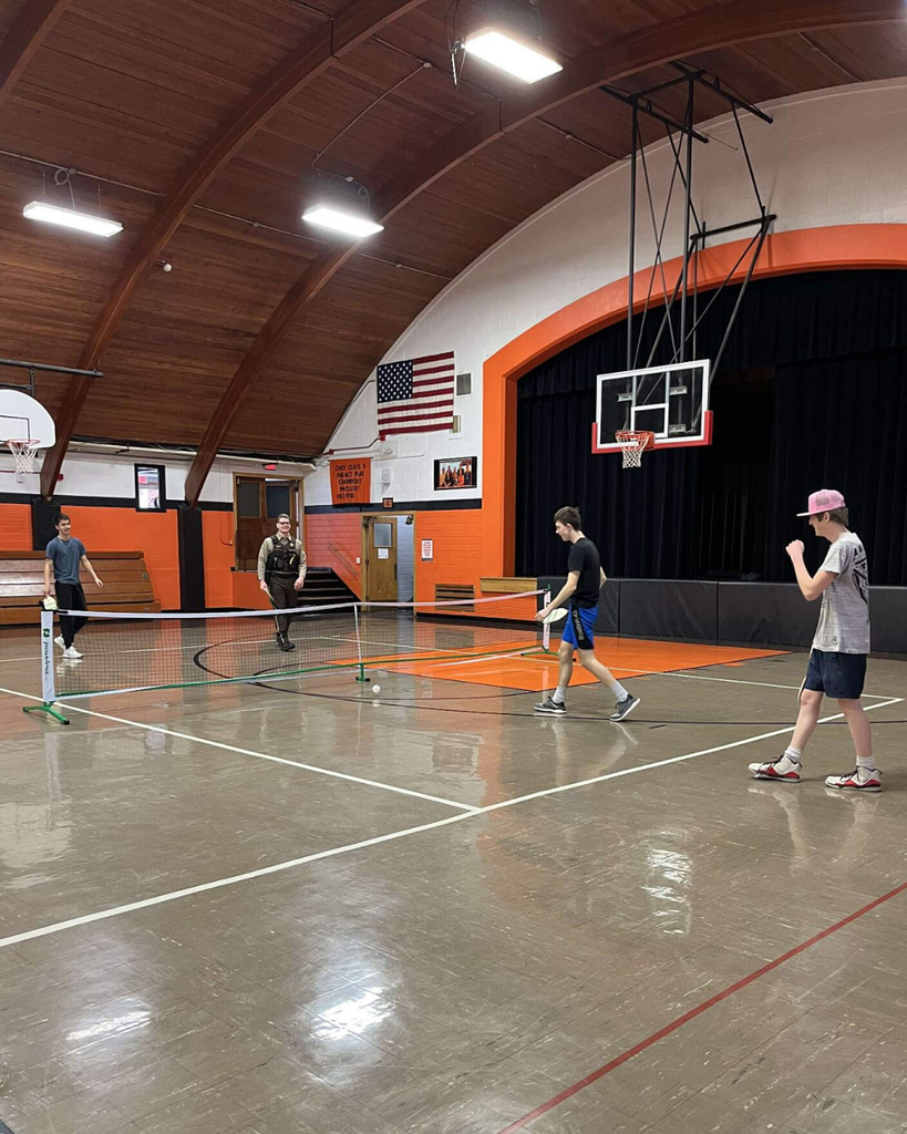A deputy sheriff playing pickleball in a high school gym with three teenage boys.