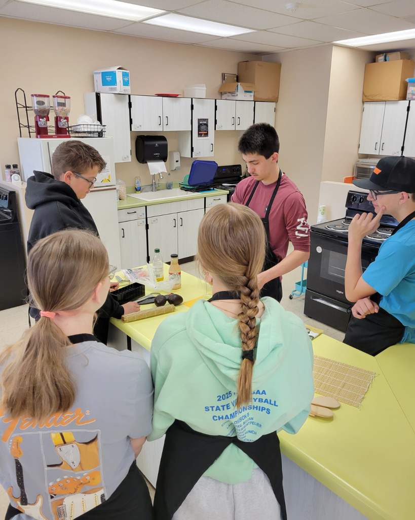 Five high school students standing around a kitchen counter watching one of them make sushi